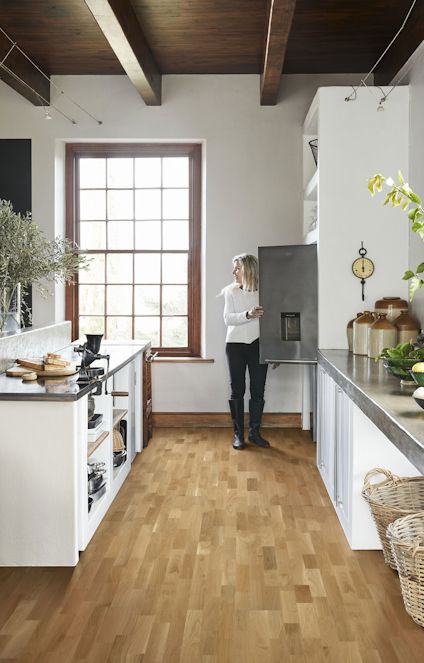 Kvinna står i ett modernt kök med trägolv och vita skåp, tittande i ett kylskåp. Stor fönsterruta mot trävägg.