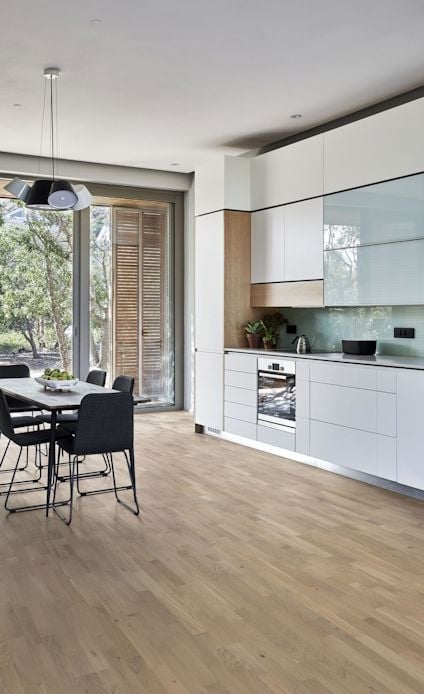 Modernt kök med ljusa trägolv, vita skåp och en glasbakvägg. Matsalsbord och stolar vid stort fönster.