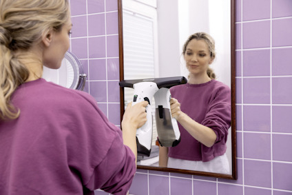 Person i lila tröja använder en elektrisk fönstertvätt på en spegel, kakelvägg i bakgrunden.